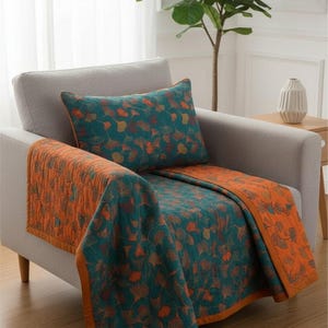 Puede incluir: Un juego de manta y cojín con estampado turquesa y naranja sobre un sillón gris. La manta y el cojín presentan un diseño de hojas repetido en turquesa, naranja y marrón. La manta está colocada sobre el brazo y el asiento de la silla.