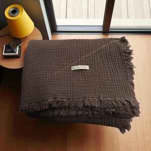 May include: A dark brown, textured blanket with fringe along the bottom edge. The blanket is folded and placed on a wooden floor. A small tag is sewn onto the blanket. A yellow lamp and small table are in the background.