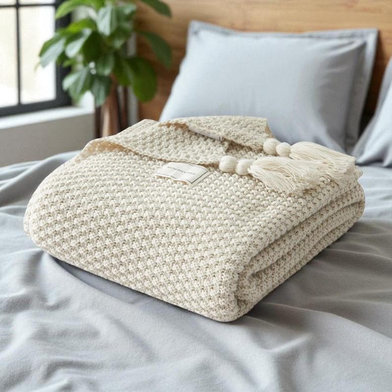 Puede incluir: Una manta tejida de color crema, con un patr&oacute;n texturizado. La manta est&aacute; adornada con borlas color crema y una peque&ntilde;a etiqueta. Se coloca sobre una cama con s&aacute;banas y almohadas grises, creando un ambiente acogedor y acogedor.