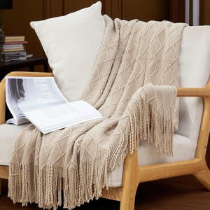 May include: A beige knitted throw blanket with a diamond pattern and fringe detail is draped over a wooden armchair. A white cushion rests on the chair, and an open book lies on the blanket. The scene suggests a cosy, comfortable living space.