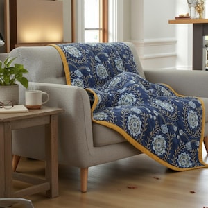 May include: A navy blue quilted throw blanket with a floral pattern in white and light blue, edged with a yellow border, draped over a gray armchair. The blanket's design features stylized flowers and leaves.