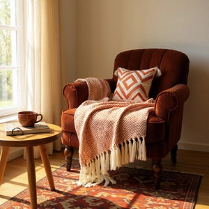 Puede incluir: Una acogedora escena de salón con un sillón color óxido con una manta estampada y un cojín decorativo. Una pequeña mesa auxiliar sostiene una taza y un libro. La alfombra y la manta comparten una paleta de colores similar.