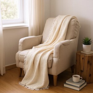 May include: A cream-colored throw blanket with fringed edges draped over a beige armchair. The chair is positioned near a window with sheer curtains. A small potted plant and a cup of coffee sit nearby, creating a cozy, inviting scene.