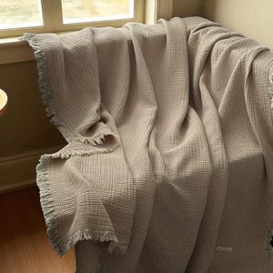 May include: A beige, textured blanket with a fringed edge is draped over a chair. The blanket has a waffle-like weave and is folded to create soft, flowing lines. The neutral color and texture suggest a cozy, home decor item.