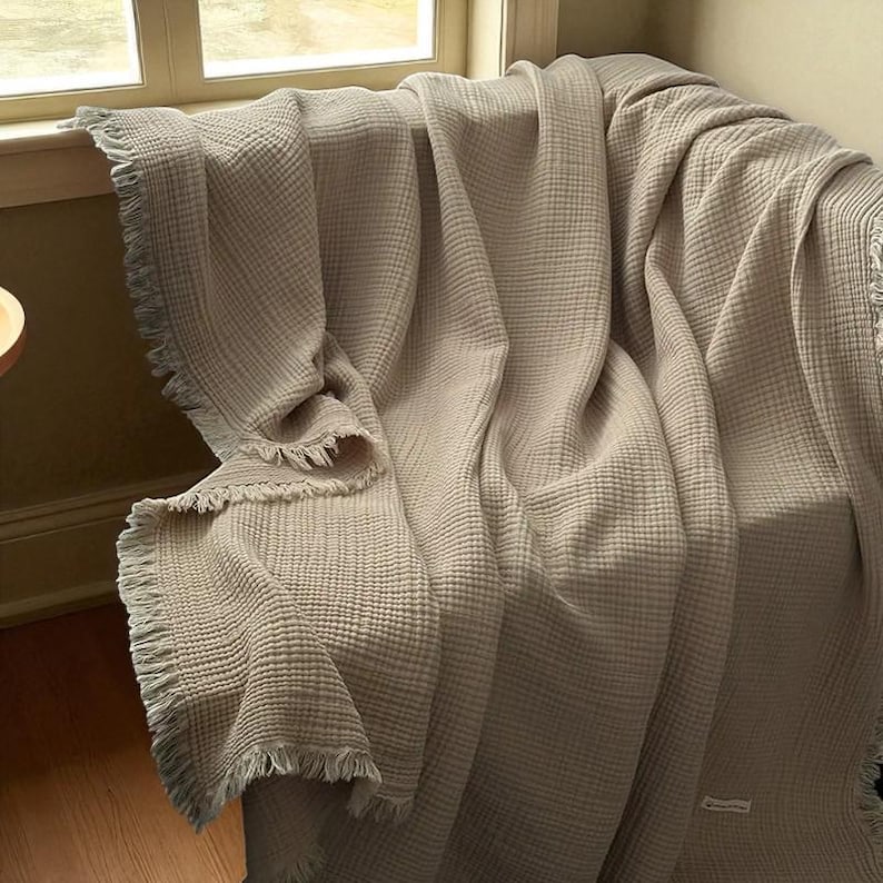 May include: A beige, textured blanket with a fringed edge is draped over a chair. The blanket has a waffle-like weave and is folded to create soft, flowing lines. The neutral color and texture suggest a cozy, home decor item.