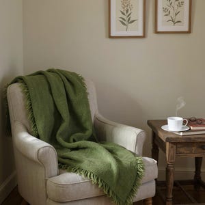 May include: A cozy living room scene featuring a light-colored armchair draped with a green fringed throw blanket. A small wooden side table holds a white teacup and saucer, with a book and glasses. Two framed botanical prints hang on the wall.