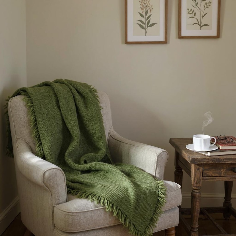 May include: A cozy living room scene featuring a light-colored armchair draped with a green fringed throw blanket. A small wooden side table holds a white teacup and saucer, with a book and glasses. Two framed botanical prints hang on the wall.