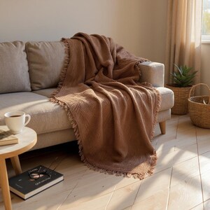 May include: A brown woven throw blanket with fringed edges draped over a beige sofa. A white mug of coffee sits on a small wooden side table next to a book and a pair of glasses. A woven basket and a potted plant are in the background.