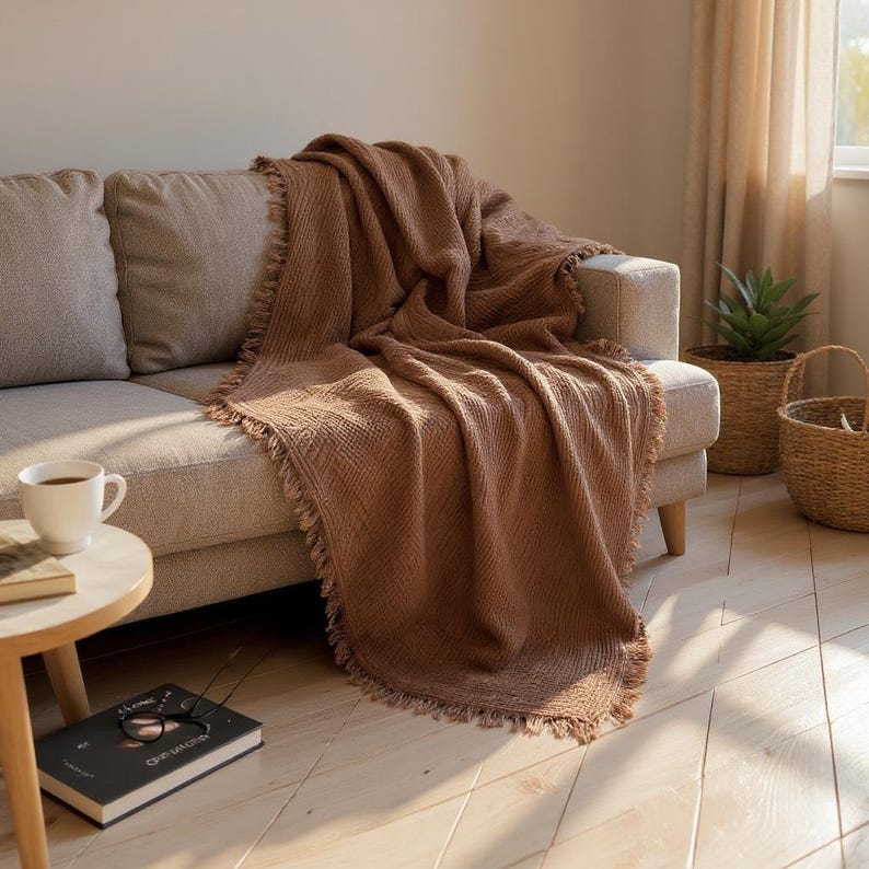 May include: A brown woven throw blanket with fringed edges draped over a beige sofa. A white mug of coffee sits on a small wooden side table next to a book and a pair of glasses. A woven basket and a potted plant are in the background.