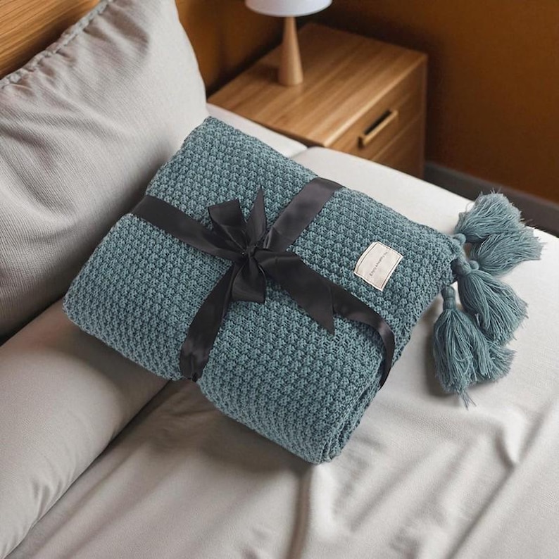 Puede incluir: Una manta de tejido de color verde azulado con un patr&oacute;n texturizado, atada con una cinta de sat&eacute;n negro. La manta tiene borlas a juego y una peque&ntilde;a etiqueta. La manta est&aacute; doblada y presentada en una cama blanca.