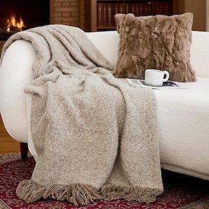 May include: A neutral-toned living room setting with a beige throw blanket and a brown fur cushion on a white sofa. A white mug rests on a magazine. A fireplace and bookcase are in the background, creating a comfortable and inviting space.