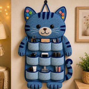 May include: A blue cat-shaped wall organizer made of crochet yarn, featuring nine individual pockets for storage. The cat has dark blue stripes, black eyes, a white muzzle, and colorful buttons on its ears. It hangs from a small black loop.
