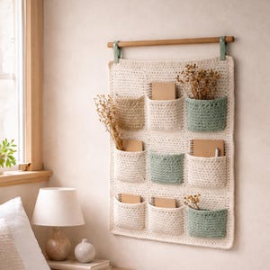 Può includere: Un organizer da parete all'uncinetto color crema con nove tasche. Le tasche sono in varie tonalità di crema e verde, e alcune contengono piccoli quaderni e fiori secchi. Un'asta di legno e un nastro verde vengono utilizzati per appenderlo.