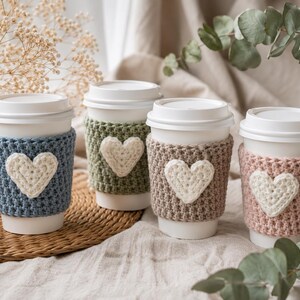 Pode incluir: Quatro copos de café descartáveis brancos, cada um com uma manga de crochê texturizada em azul, verde sálvia, bege ou rosa seco. Cada manga é adornada com uma aplicação de coração de crochê na cor creme centralizada.