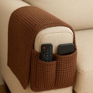 Könnte beinhalten: Ein brauner, gehäkelter Sofa-Armlehnen-Organizer mit zwei Taschen. Eine Tasche enthält eine schwarze Fernbedienung, die andere ein schwarzes Smartphone. Der Organizer ist über der Armlehne eines beigen Sofas drapiert.