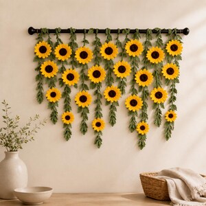 May include: A decorative wall hanging featuring yellow sunflowers with dark brown centers and green leaves. The sunflowers are attached to a black rod, creating a vibrant and natural-looking floral display. The arrangement adds a touch of nature to any space.