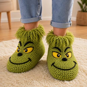 Puede incluir: Un par de zapatillas de ganchillo verdes con una cara de personaje, ojos amarillos, cejas negras y una sonrisa cosida en negro. Las zapatillas tienen un borde de flecos verdes alrededor del tobillo y un aspecto tejido a mano.