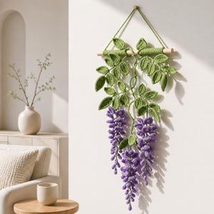 Può includere: Un arazzo da parete all'uncinetto fatto a mano con foglie verdi e fiori di glicine viola. Il design floreale è sospeso da un tassello di legno e un cordino verde, aggiungendo un tocco di decorazione ispirata alla natura.