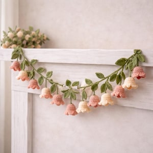 Op de afbeelding: Een handgemaakte gehaakte slinger met roze en crèmekleurige bloemknoppen en groene bladeren. De bloemenversiering is geregen langs een groene slinger, perfect om een vleugje lente aan een ruimte toe te voegen.