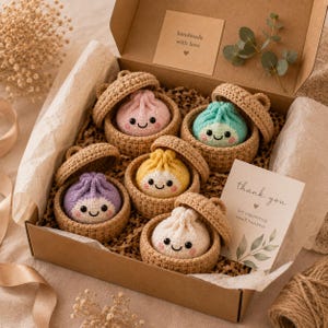 Peut inclure: Une boîte cadeau en carton contenant cinq jouets en forme de raviolis au crochet, chacun niché dans un petit panier en crochet beige avec couvercle. Les raviolis sont roses, bleu sarcelle, jaunes, violets et blancs, avec des visages souriants.
