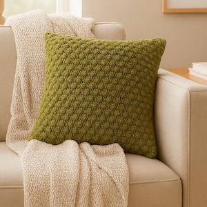 May include: An olive green cushion with a textured, diamond pattern sits on a beige sofa. A cream-coloured, textured throw blanket is draped over the sofa. The scene suggests a comfortable, inviting living space. Home decor.