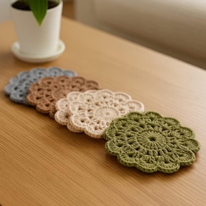 Op de afbeelding: Een rij van vijf handgemaakte gehaakte onderzetters, elk in een andere kleur. De kleuren zijn grijs, bruin, beige en olijfgroen. De onderzetters zijn cirkelvormig met een geschulpte rand en een gedetailleerd open patroon.