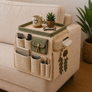 May include: A cream-coloured crocheted sofa arm organiser with various pockets for storage. It has a green trim, a small green pocket with a button, and a sunflower appliqué. Includes a tray for a mug and a small plant.