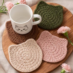 Könnte beinhalten: Eine weiße Keramiktasse mit Katzengesicht-Design steht auf einem braunen, gehäkelten, katzenförmigen Untersetzer. Drei weitere katzenförmige Untersetzer in Grün, Rosa und Beige umgeben die Tasse. Die Untersetzer befinden sich auf einer Holzoberfläche, mit rosa Blumen.