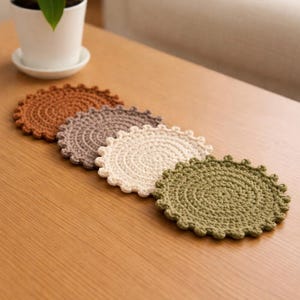 May include: Five handmade crochet coasters in shades of brown, gray, cream, and green are arranged on a wooden surface. Each coaster has a circular shape with a scalloped edge, showcasing a detailed, textured design. The coasters are a decorative and functional home accessory.