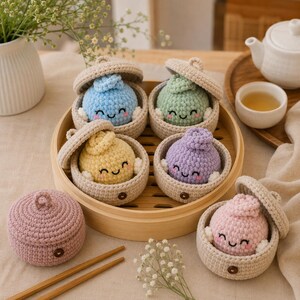 Puede incluir: Un conjunto de seis recipientes de ganchillo con forma de dumpling en tonos pastel azul, verde, amarillo, morado y rosa, cada uno con tapa a juego y un pequeño botón marrón en el lateral. Los recipientes tienen caras sonrientes bordadas en una vaporera.