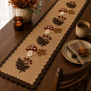 May include: A rectangular crochet table runner in a tan hue with a dark brown scalloped border. The runner features repeating appliqués of red and white mushrooms, dark brown fern-like leaves, and small berry clusters arranged in a line.