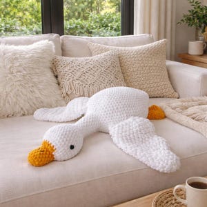 Könnte beinhalten: Ein großes, weißes, gehäkeltes gänseförmiges Kissen mit gelbem Schnabel und Füßen liegt auf einem cremefarbenen Sofa. Das Kissen ist von anderen dekorativen Kissen in verschiedenen Texturen und Cremetönen umgeben, was eine gemütliche, einladende Atmosphäre schafft.