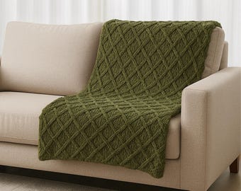 May include: A green knitted throw blanket with a diamond pattern is draped over the back of a beige sofa. The blanket's texture and color provide a cozy and inviting aesthetic. The sofa has a simple, modern design.