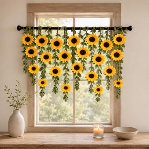 May include: A decorative window treatment featuring yellow sunflowers with dark brown centers and green vines. The sunflowers are arranged in a cascading pattern, hanging in front of a window. A vase, candle, and bowl are on the wooden windowsill.