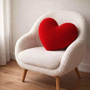 May include: A red heart-shaped pillow rests on a white, textured armchair with wooden legs. The pillow is the focal point, its vibrant color contrasting with the neutral tones of the chair and background. The chair has a rounded back and armrests.