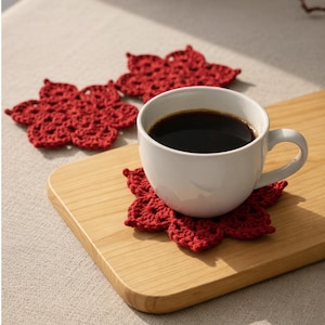 Puede incluir: Una taza de cerámica blanca llena de café oscuro se encuentra sobre un posavasos rojo de ganchillo con forma de flor. Dos posavasos de flores rojas adicionales están sobre una tabla de cortar de madera. Los posavasos son hechos a mano.