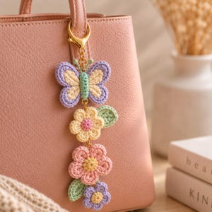 Op de afbeelding: Een gehaakte vlinder- en bloemenbedel in pasteltinten, opgehangen aan een gouden sluiting die aan een roze handtas is bevestigd. De vlinder heeft lavendel-, gele en groene details. De bloemen zijn geel, roze en lavendel.
