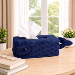 May include: A navy blue crocheted tissue box cover shaped like a whale, with a smiling face and a tail. White tissues are pulled from the top opening. The cover is on a wooden table, with a pair of glasses and books in the foreground.