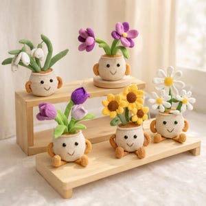May include: A selection of crocheted flower pots, each with a smiling face and holding a different type of flower. The flowers include snowdrops, purple tulips, sunflowers, daisies, and purple flowers. The pots are beige with orange arms and legs.