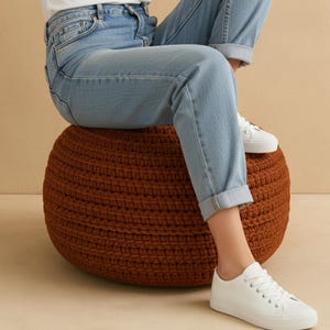 Puede incluir: Un otomán redondo de punto marrón. El otomán tiene un tono cálido y terroso y una superficie texturizada. Zapatillas blancas son visibles en el otomán y en el suelo. El otomán es un mueble decorativo.