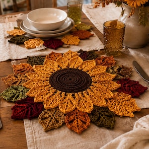 May include: A round, crocheted placemat featuring a dark brown center surrounded by layers of yellow, orange, red, and olive green leaf-shaped petals. The intricate, open-work design mimics the appearance of a sunflower with autumn-colored foliage.