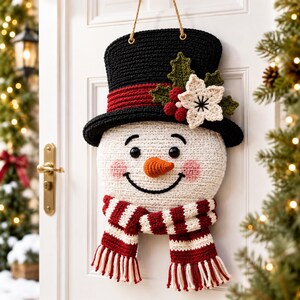 Peut inclure: Une d&eacute;coration de porte en crochet repr&eacute;sentant un bonhomme de neige avec un haut-de-forme noir &agrave; bande rouge, des feuilles vertes, des baies rouges et une fleur blanche. Il a un visage blanc, des yeux noirs, un nez orange et une &eacute;charpe ray&eacute;e.