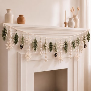 Op de afbeelding: Een decoratieve slinger met gehaakte elementen, waaronder groene eikenbladeren, witte sneeuwvlokken, kleine witte bessenclusters en bruine dennenappels. De items hangen aan een lang, crèmekleurig gehaakt koord in een herhalend patroon.