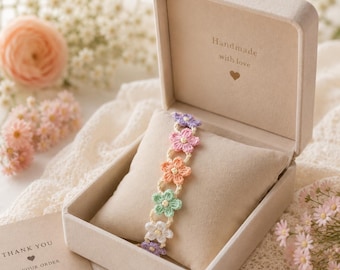Può includere: Un braccialetto fatto a mano con fiori all'uncinetto nei colori pastello viola, rosa, arancione, verde e bianco. Il braccialetto è esposto in un portagioie beige aperto con la scritta "Handmade with love".