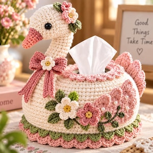 May include: A cream-colored crochet tissue box cover shaped like a swan with a pink beak, a pink bow at the neck, and floral embellishments. The swan features green leaves, white and pink flowers, and small white pearls on the wings.