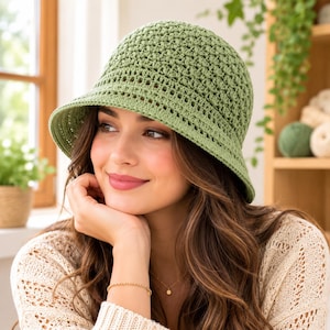 May include: A sage green crochet bucket hat featuring a textured, dense stitch pattern on the crown and a decorative, open-work band detail above the brim. The hat is constructed from yarn and has a rounded shape with a downward-sloping brim.