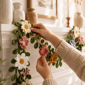 May include: A decorative garland featuring crocheted flowers in white, pink, and orange, interspersed with green crocheted leaves. The floral arrangement is crafted with intricate yarn textures and is displayed draped over a white mantelpiece.