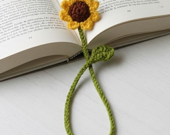 Puede incluir: Marcapáginas de ganchillo hecho a mano con forma de girasol. El girasol tiene pétalos amarillos y un centro marrón. El tallo y la hoja son verdes. El marcapáginas está en un libro abierto.