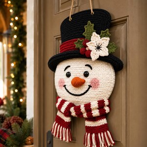 Peut inclure: Une d&eacute;coration murale en crochet repr&eacute;sentant un bonhomme de neige avec un haut-de-forme noir &agrave; bande rouge, orn&eacute; de feuilles de houx vertes, de baies rouges et d'une fleur blanche. Il poss&egrave;de un visage blanc, des yeux noirs et une &eacute;charpe ray&eacute;e.