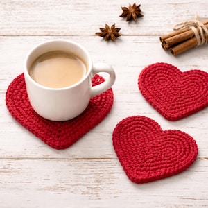 May include: A white mug filled with coffee sits on a red heart-shaped crochet coaster. Two additional red heart coasters are visible, along with star anise and cinnamon sticks, creating a cozy, romantic theme.